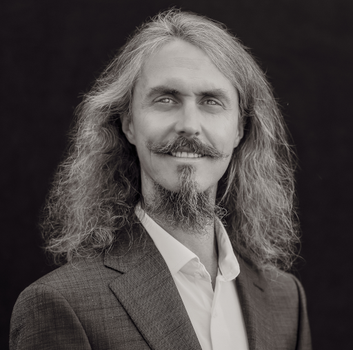 A black and white photo of a white man with long hair, mustache and goatee, wearing a suit jacket with white dress shirt.